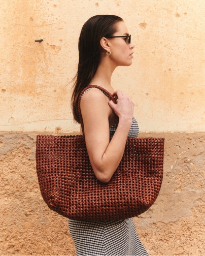 Side profile of woman with slicked back dark hair and sunglasses in front of a stone wall wearing a leather bag on her shoulder